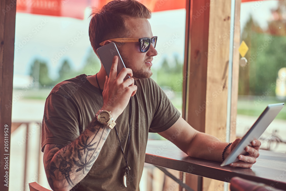 Handsome stylish hipster sits at a table in a roadside cafe, talking on the phone and holds the tablet. Handsome stylish hipster sits at a table in a roadside cafe, talking on the phone and holds the tablet.