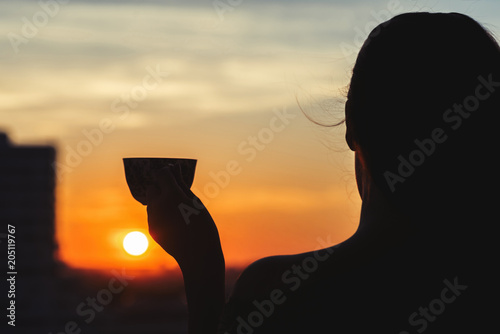  Silhouette of girl with a cup of tea at sunset. View on the town from above