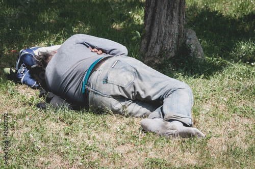 Old barefoot homeless or refugee man sleeping on the grass in the park using his travel bag as pillow, social documentary street concept