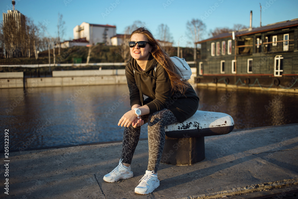 Urban style and fashion concept. Outdoor portrait of beautiful stylish young European female model with long brown hair wearing trendy hoodie, sunglasses, white sneackers and white watches, posing