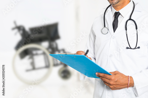 Doctor with stethoscope and clipboard writing a medical prescription with medical wheelchair in background