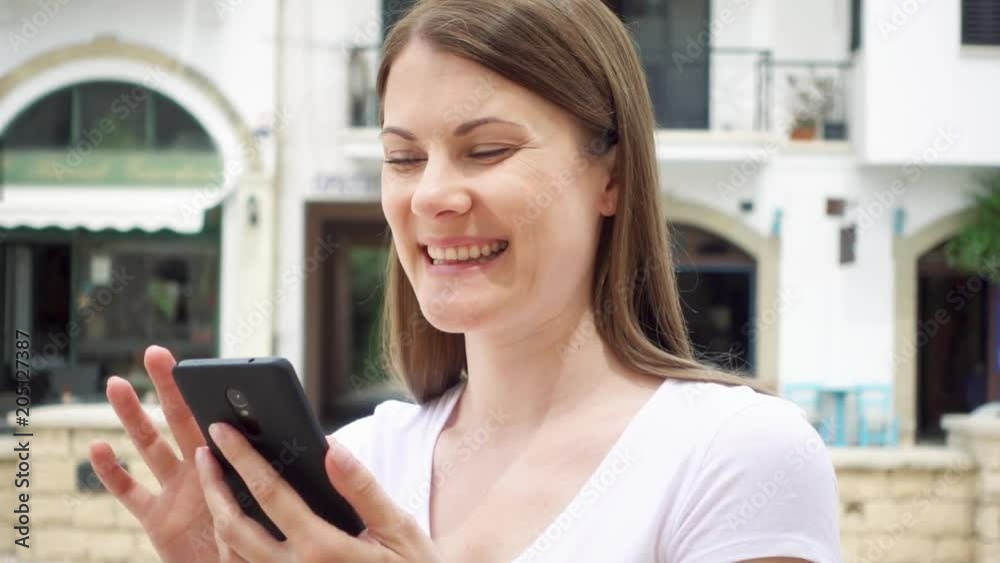 Smiling young woman in white t-shirt standing on street in european city browsing on cellphone in slow motion. Happy female chatting with friends via mobile phone Smiling young woman in white t-shirt standing on street in european city browsing on cellphone in slow motion. Happy female chatting with friends via mobile phone