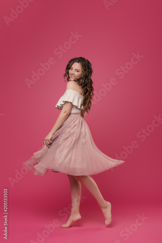 Young stylish girl in pink light skirt and white T-shirt on pink isolated background with place for text. The girl cheerfully dances and jumps like a ballerina