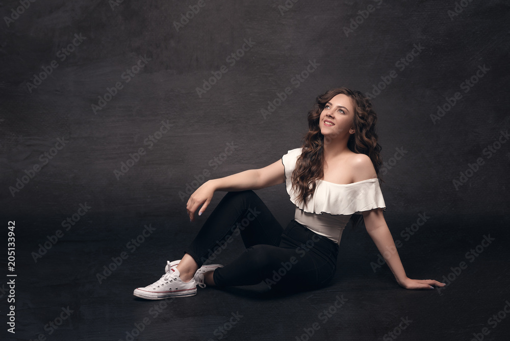 Young stylish girl model with long curly hair in stylish clothes sits on a dark isolated background with space for text