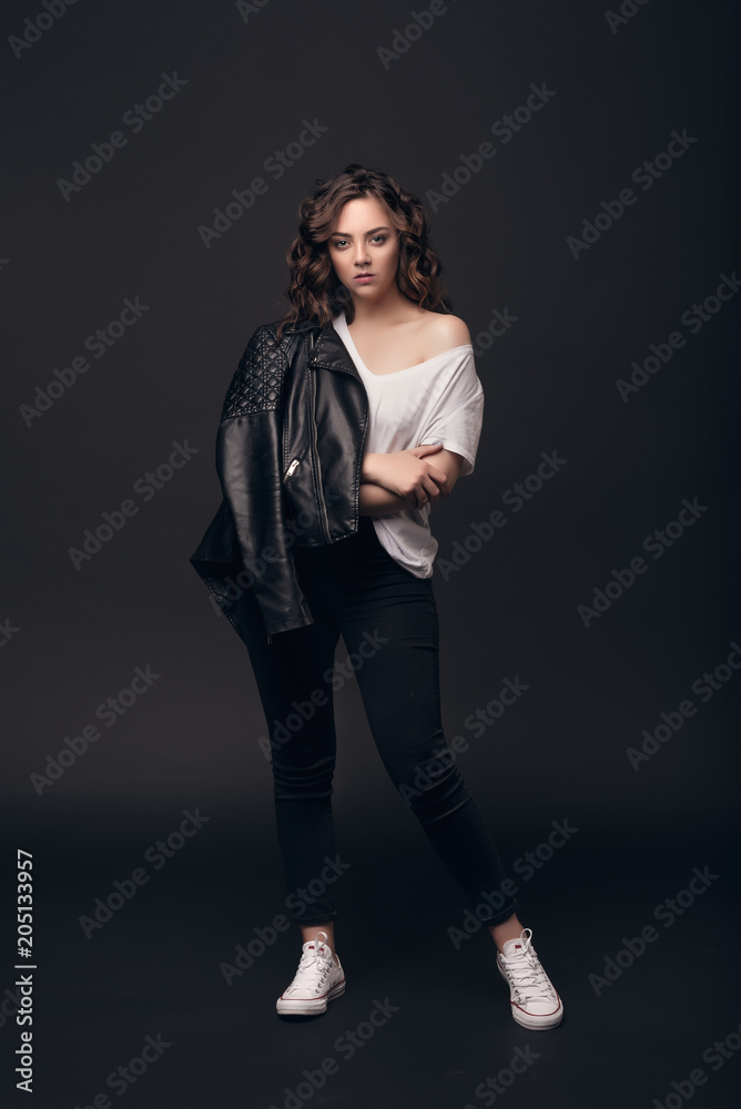 A young stylish girl model with long curly hair in a stylish black jacket, jeans, white sneakers and a white T-shirt is standing on a dark, isolated background in the studio. A young stylish girl model with long curly hair in a stylish black jacket, jeans, white sneakers and a white T-shirt is standing on a dark, isolated background in the studio.