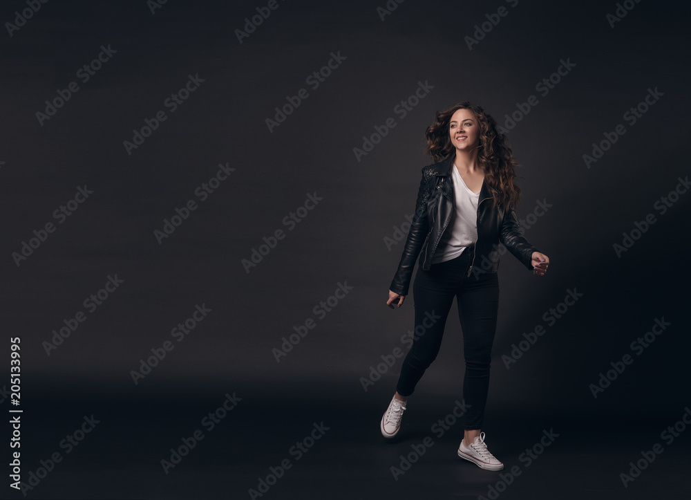 Young stylish girl model with long curly hair in a stylish black jacket, jeans, white sneakers joyfully runs on a dark isolated background. Place for textt
