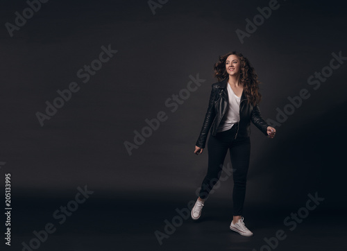 Young stylish girl model with long curly hair in a stylish black jacket, jeans, white sneakers joyfully runs on a dark isolated background. Place for textt