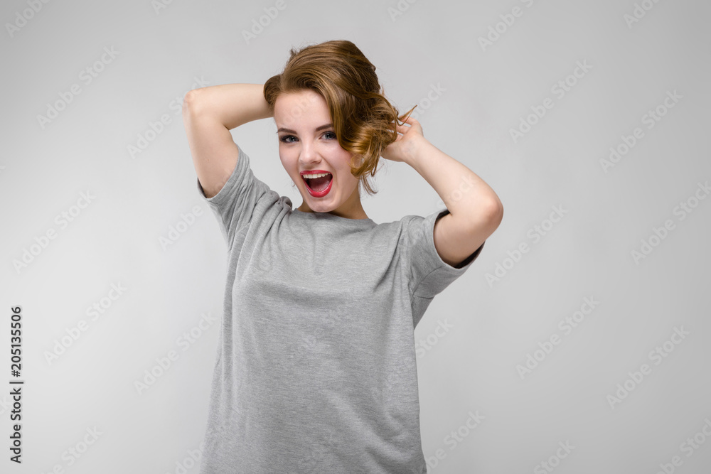 Charming young girl in a gray T-shirt on a gray background. The girl holds her hands behind her head and screams Charming young girl in a gray T-shirt on a gray background. The girl holds her hands behind her head and screams