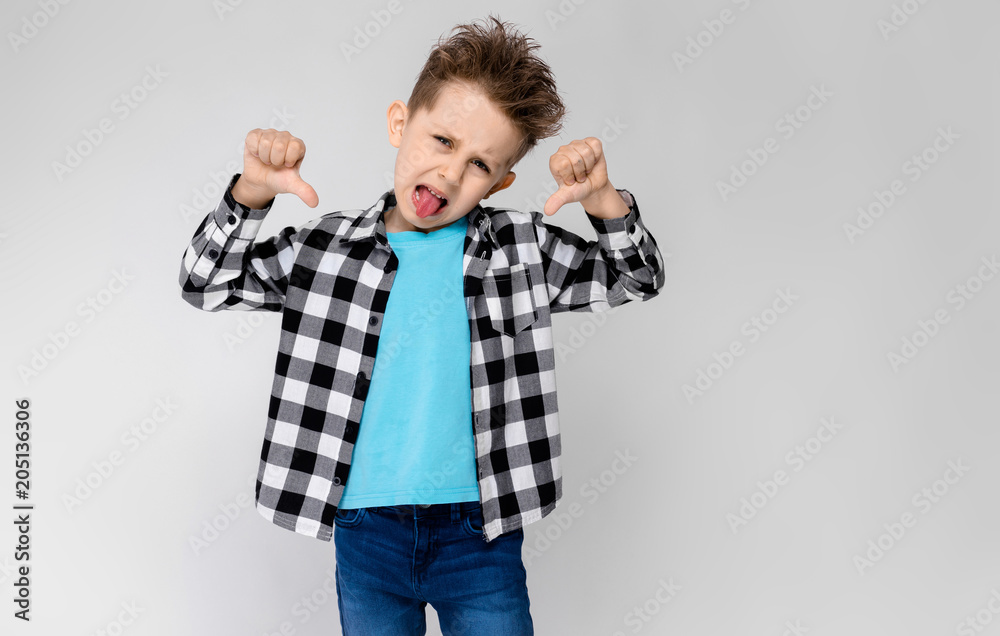 A handsome boy in a plaid shirt, blue shirt and jeans stands on a gray background. The boy folded his arms over his chest. Boy showing thumb down A handsome boy in a plaid shirt, blue shirt and jeans stands on a gray background. The boy folded his arms over his chest. Boy showing thumb down
