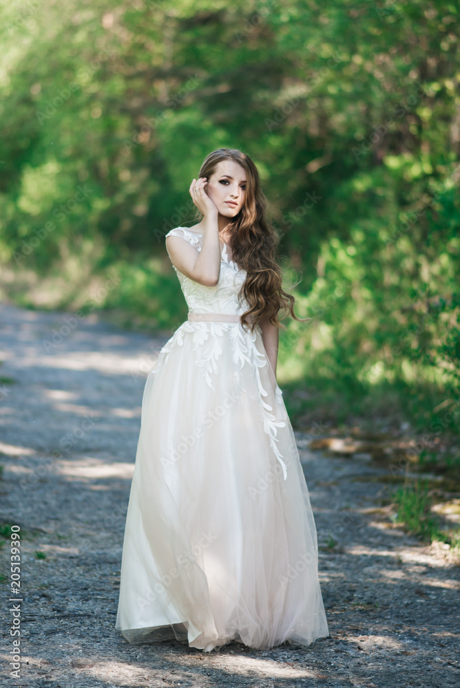 Beautiful brunette bride with curls, makeup and stylish dress. Portrait on the background of green trees. Summer wedding day.
