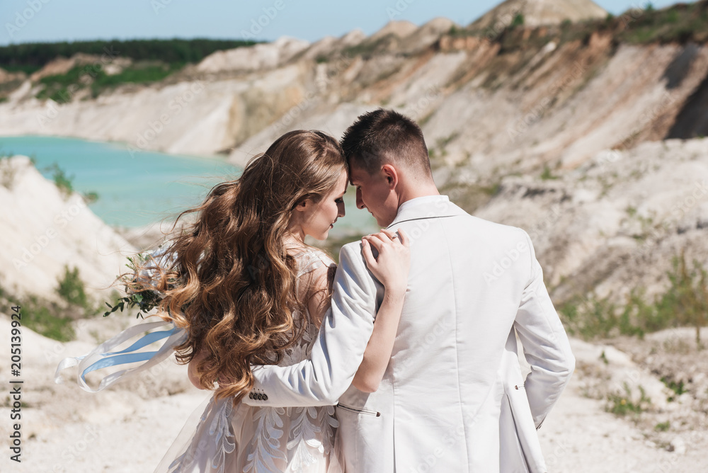 The bride in a beautiful dress hugging the groom in a light suit near the lake. Wedding couple standing on a sandy hill in the open air. A romantic love story. Azure-blue water on the horizon. The bride in a beautiful dress hugging the groom in a light suit near the lake. Wedding couple standing on a sandy hill in the open air. A romantic love story. Azure-blue water on the horizon.