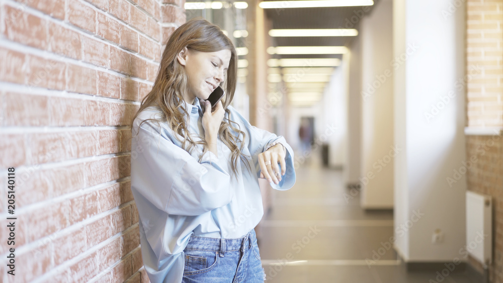 Young woman wearing a white shirt and jeans is talking on her smartphone and looking on her watch standing in a business building corridor.