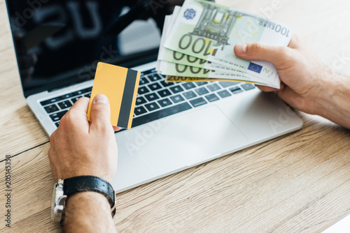 cropped shot of person holding credit card and euro banknotes above laptop