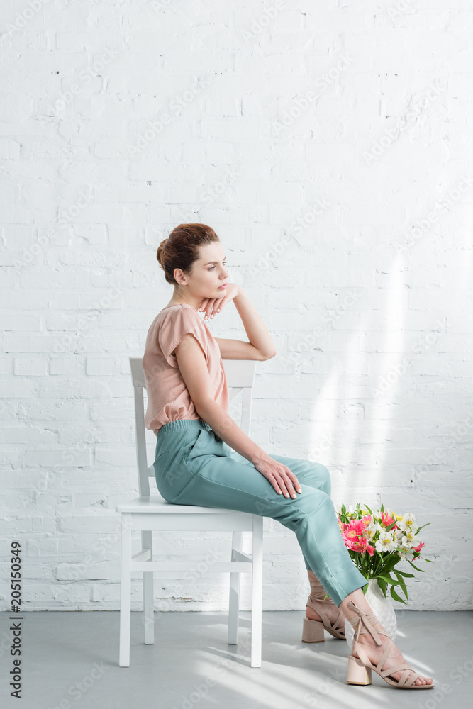 thoughtful young woman sitting on chair with flowers on floor in front of white brick wall
