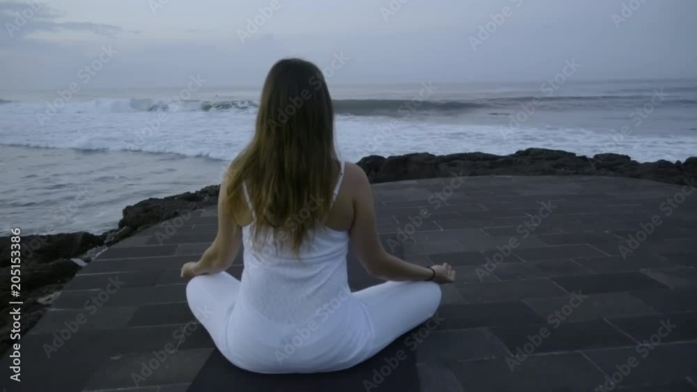 Zoom in with rear view of woman in white outfit sitting in lotus pose on stone dock near ocean and meditating in morning Zoom in with rear view of woman in white outfit sitting in lotus pose on stone dock near ocean and meditating in morning