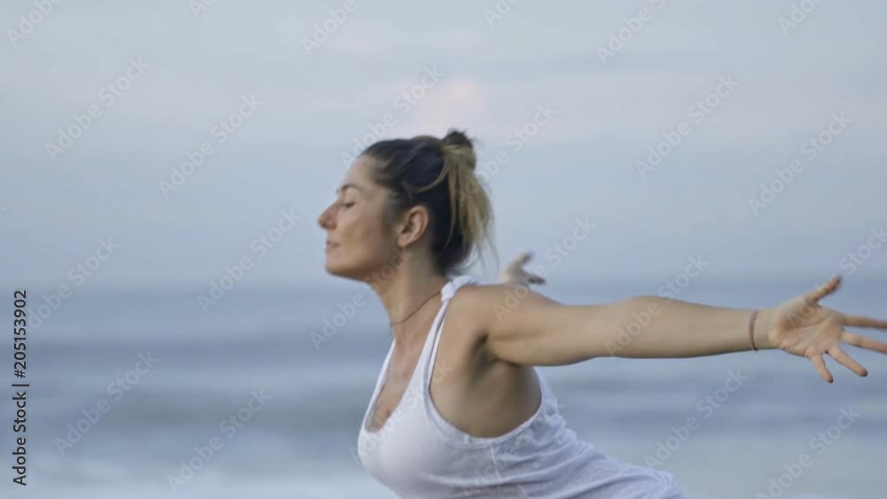 Medium shot of mature woman in white doing yoga on beach: she performing raised arms pose, then transitioning to forward bend Medium shot of mature woman in white doing yoga on beach: she performing raised arms pose, then transitioning to forward bend