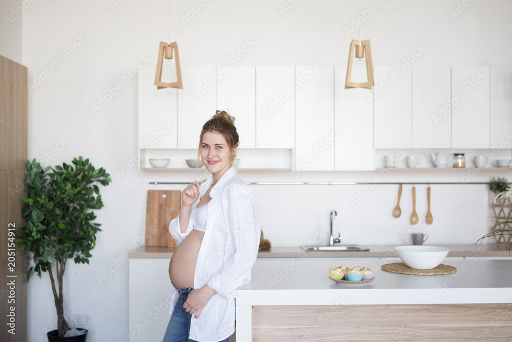 A pregnant woman is standing in the kitchen in jeans and a white shirt A pregnant woman is standing in the kitchen in jeans and a white shirt