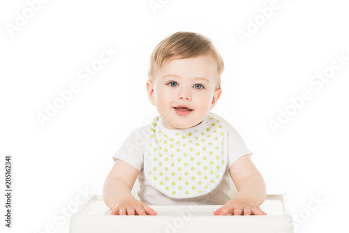 smiling baby boy in bib sitting in highchair isolated on white background