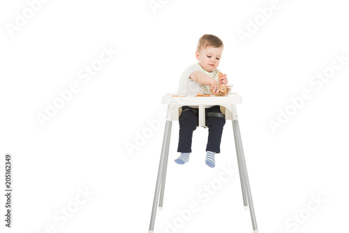 little boy in bib eating bagels and sitting in highchair isolated on white background