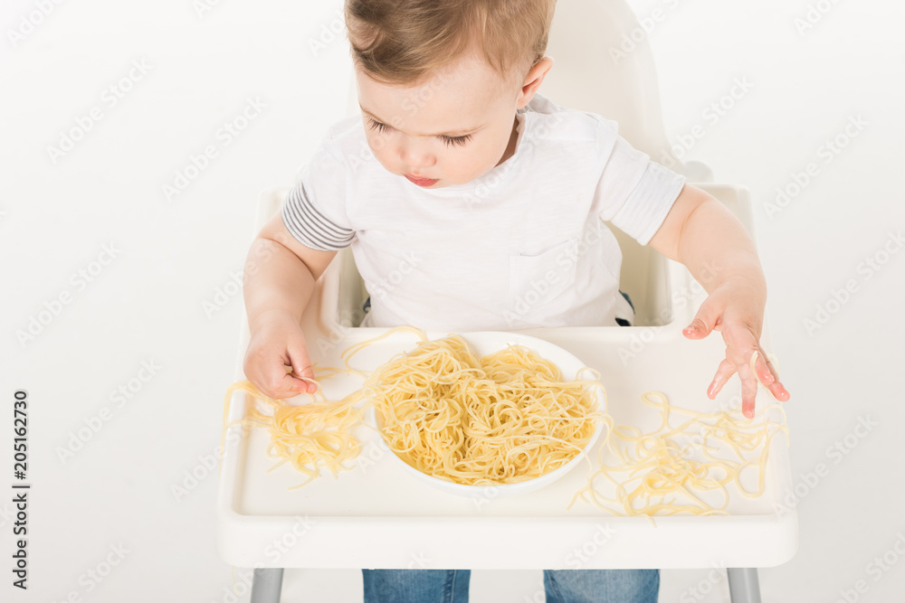 high angle view of baby boy sitting in highchair and eating spaghetti by hands high angle view of baby boy sitting in highchair and eating spaghetti by hands