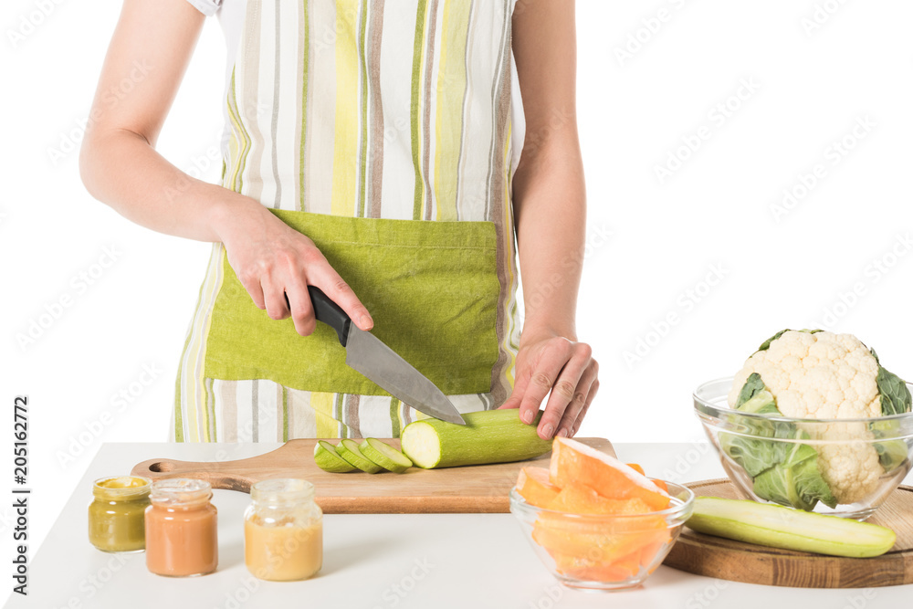 cropped shot of woman cutting zucchini by knife at table with puree jars, cauliflower and pumpkin cropped shot of woman cutting zucchini by knife at table with puree jars, cauliflower and pumpkin