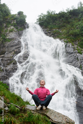 Happy woman sitting in lotus position near waterfall meditating with her eyes closed copyspace