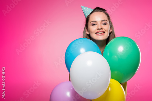 attractive woman with party hat looking out from balloons isolated on pink