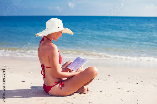 Caucasian woman is reading a book on a sandy beach wearing a straw hat