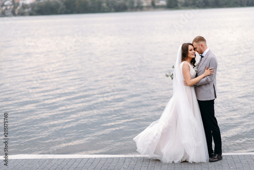 An amazing couple on their wedding day near the lake hug and enjoy each other. The bride and groom with a bouquet are happy together