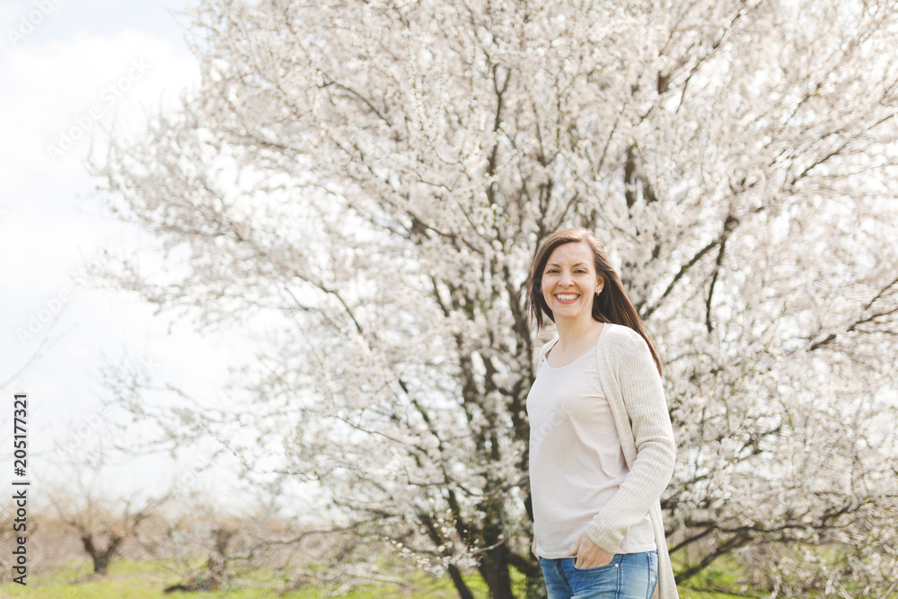 Young happy smiling beautiful woman in light casual clothes keeping hands in pockets standing in city garden or park on blooming tree background. Spring nature, flowers. Lifestyle, leisure concept. Young happy smiling beautiful woman in light casual clothes keeping hands in pockets standing in city garden or park on blooming tree background. Spring nature, flowers. Lifestyle, leisure concept.