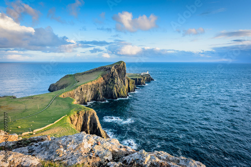 Neist Point lighthouse at sunset on a bright sunny day with clear waters, dramatic sky, gentle rays of sunlight over green grass and jagged rocks - typical Scottish landscape