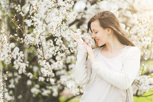 Young relaxed tender beautiful woman with closed eyes in light casual clothes sniffing flowers in city garden or park on blooming tree background. Spring nature, flowers. Lifestyle, leisure concept.