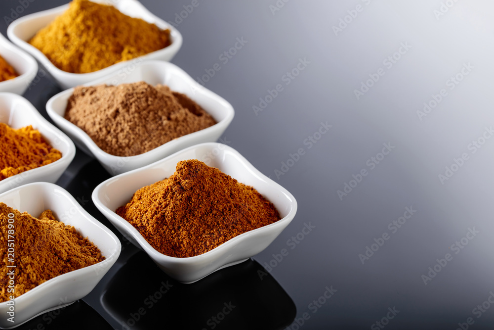Small white ceramic bowls of Indian spices on black reflective background.