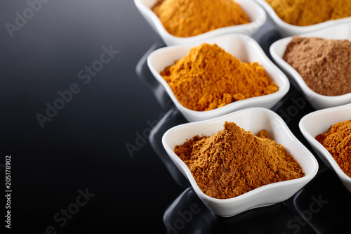 Small white ceramic bowls of Indian spices on black reflective background.