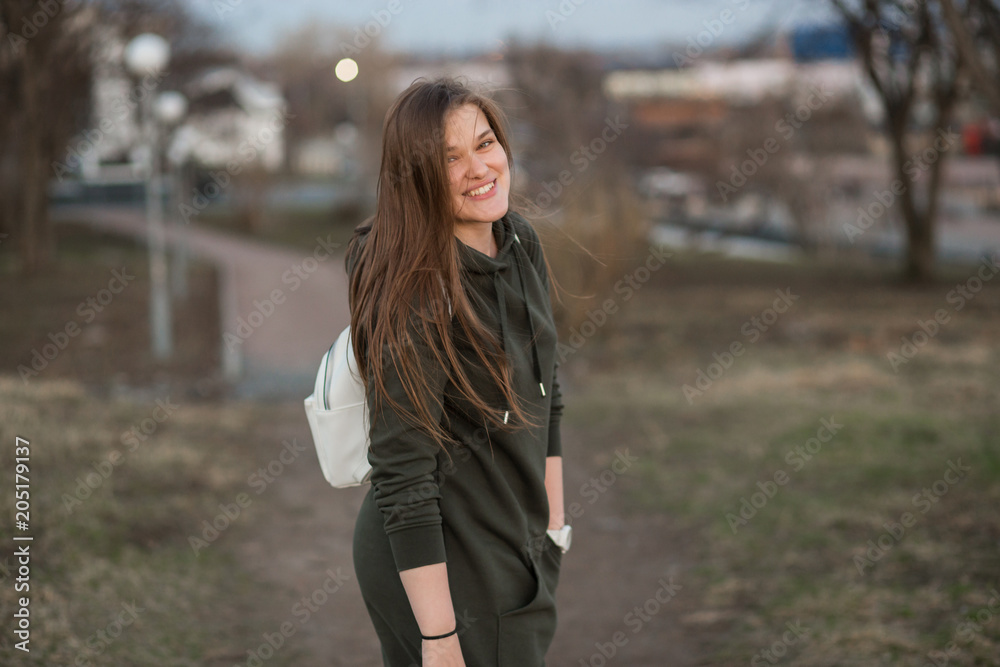 Outdoor portrait of beautiful stylish young female model with long brown hair wearing trendy hoodie and white walking in evening autumn or spring park