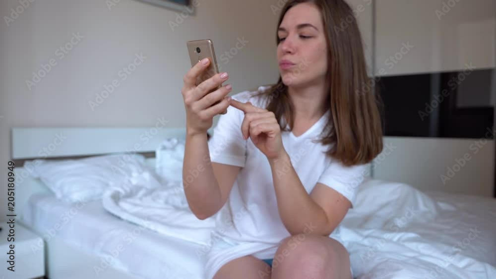 Young Pretty Woman Makes Selfie On A Smartphone On The Background Of Bed Prevents Sleep To Blurred Angry Husband Young Pretty Woman Makes Selfie On A Smartphone On The Background Of Bed Prevents Sleep To Blurred Angry Husband