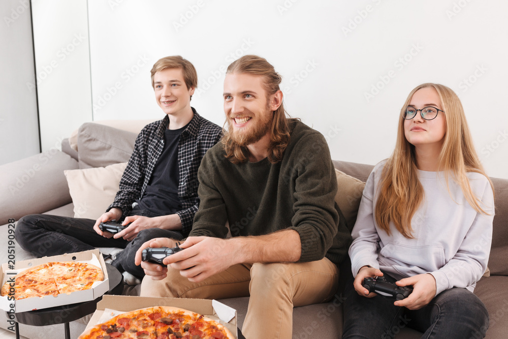 Group of cheerful friends sitting on sofa and playing video games together at home. Portrait of two boys and girl sitting with joysticks and pizza isolated Group of cheerful friends sitting on sofa and playing video games together at home. Portrait of two boys and girl sitting with joysticks and pizza isolated