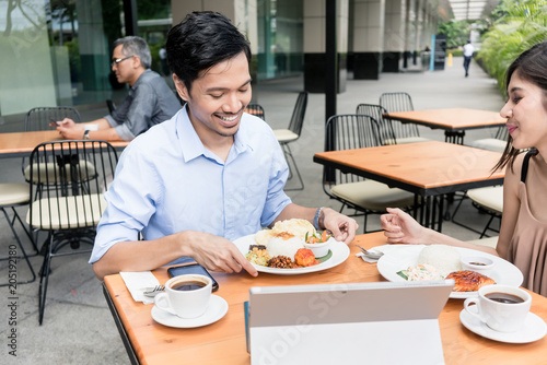 Young Asian man and woman smiling while having lunch together outdoors at a modern cafeteria downtown in summer