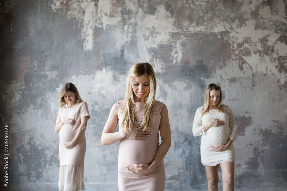 Three pregnant women stand with happy smiles and look at the belly.One pregnant woman in focus and two pregnant women in a defocus. Back background gray wall
