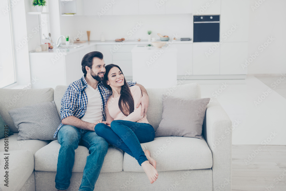 Obraz premium Portrait of attractive cheerful couple spending time together indoor, in house, apartment sitting on sofa looking to the side. True feelings harmony idyllic affection concept