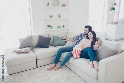 Portrait of trendy attractive couple sitting in modern living room using console trying to find interesting channel program film on television hugging embracing eating snack
