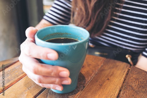 Closeup image of a woman's hand holding blue mug of hot coffee on vintage wooden table in cafe