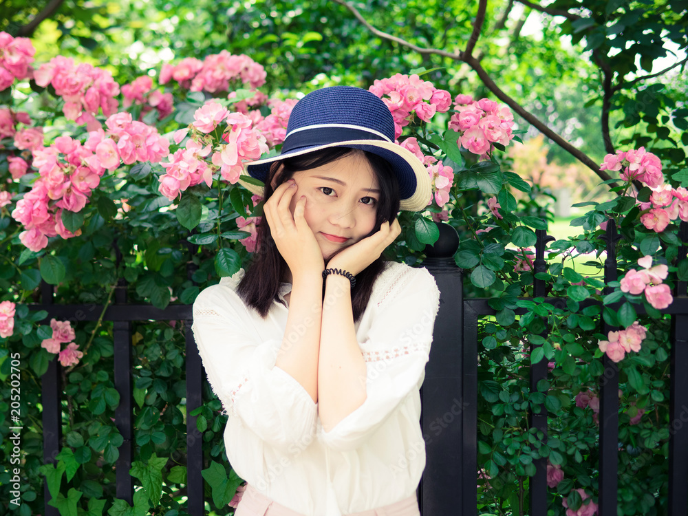 Outdoor portrait of beautiful young Chinese woman in casual dress smiling among pink rose flowers wall in spring garden.