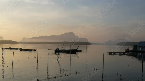 silhouette fisherman sail long tail boat around  a floating basket for keeping live fish in brine sea water. Fog , silhouette mountain, mangrove tree forest, blue sky are background. 