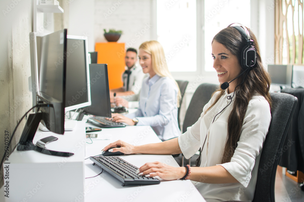 Portrait of beautiful and cheerful young woman telephone operator with headset working on desktop computer in row in customer service call support helpline business center with co-workers Portrait of beautiful and cheerful young woman telephone operator with headset working on desktop computer in row in customer service call support helpline business center with co-workers