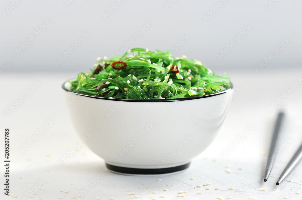 Salad chuka from seaweed in a white bowl. Salad sprinkled with sesame seeds. In the frame, chopsticks. Light background. Close-up. Macro photography. Salad chuka from seaweed in a white bowl. Salad sprinkled with sesame seeds. In the frame, chopsticks. Light background. Close-up. Macro photography.