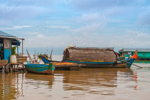 Photography A wooden sampan-like boat with a curved roof tied up to a floating house in Chong Kneas, the floating village on Tonle Sap Lake in Cambodia