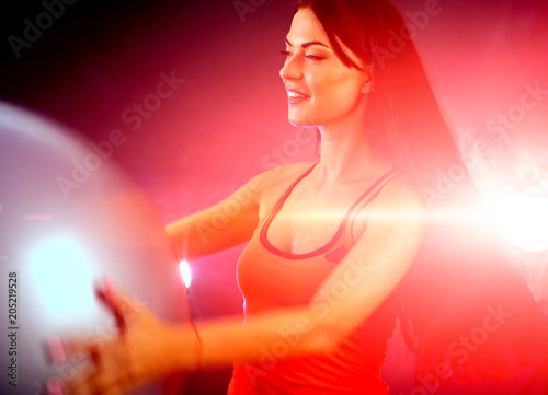 Fitness girl exercising in gym with ball. Woman with fitball works in multi-colored rays at aerobics of sport room. Portrait of full-length in profile with lens flare. Dreams of slender body.