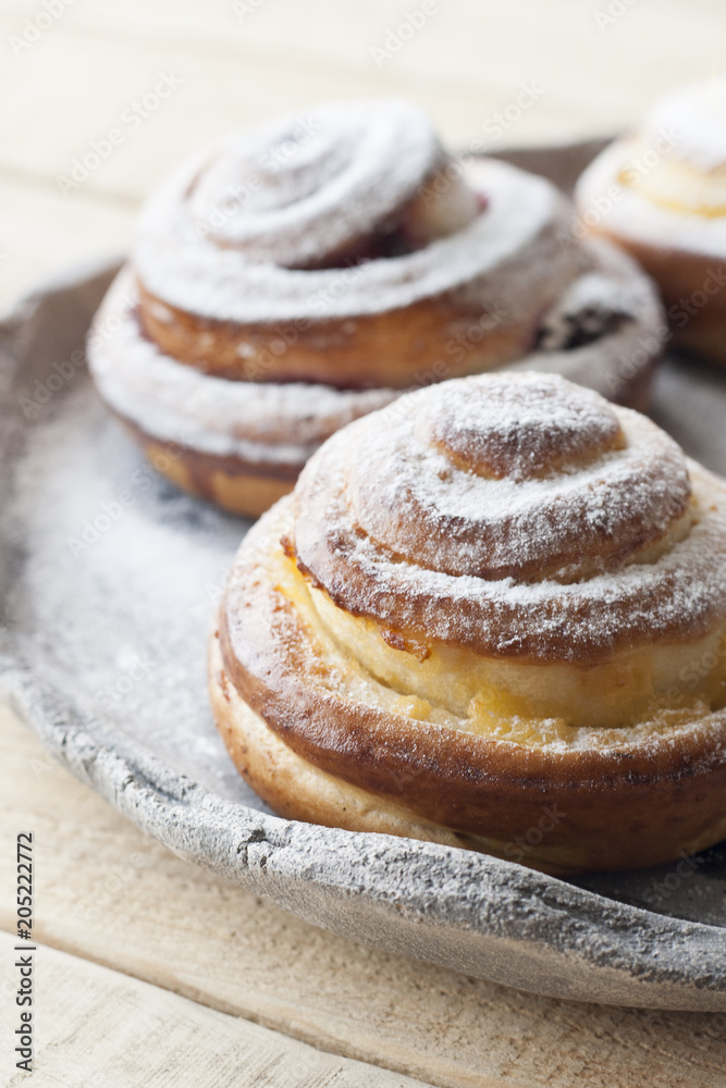 Tasty buns are sprinkled with sugar powder on a light hand-made plate on a light wooden background.