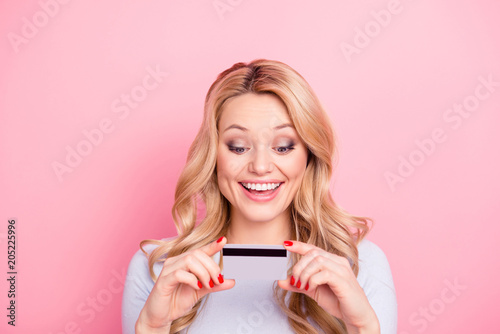 Portrait of glad excited girl with modern curly hairdo looking at credit card in her hands going to make online shopping spending a lot of money isolated on pink background